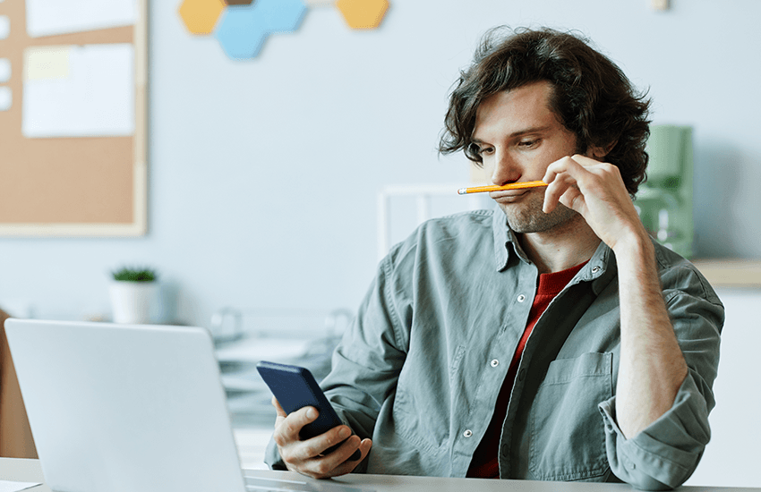Junger Mann sitzt am Schreibtisch, hält ein Smartphone und balanciert gedankenverloren einen Stift unter der Nase – Symbol für Konzentrationsprobleme und Ablenkung im Arbeitsalltag.