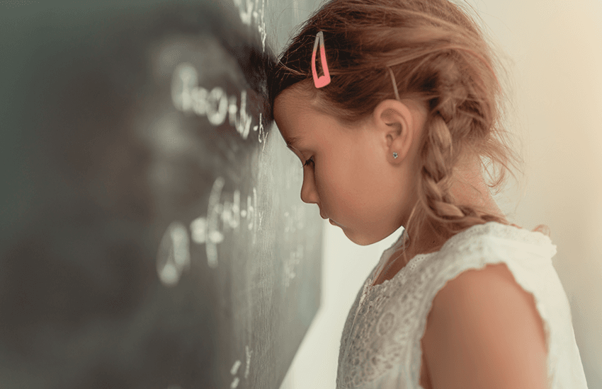 Mädchen mit geflochtenen Haaren lehnt frustriert die Stirn gegen eine Tafel mit Mathematikformeln – Ausdruck von Überforderung oder Konzentrationsproblemen in der Schule.