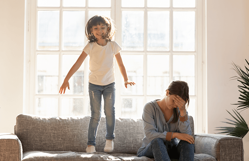 Kleines Mädchen springt energiegeladen auf einem Sofa, während die Mutter daneben sitzt, den Kopf gestützt und erschöpft wirkt – Symbol für Überforderung durch kindliche Hyperaktivität durch ADHS.