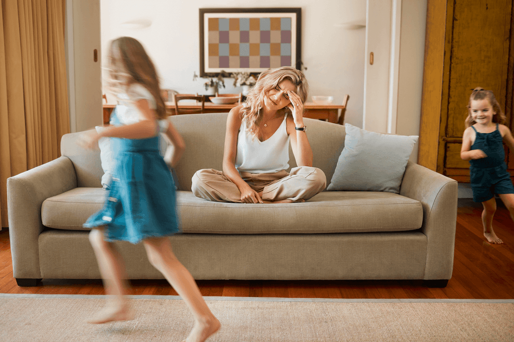 Zwei kleine Mädchen rennen ausgelassen durch ein Wohnzimmer, während ihre Mutter erschöpft mit geschlossenen Augen und einer Hand an der Stirn auf dem Sofa sitzt