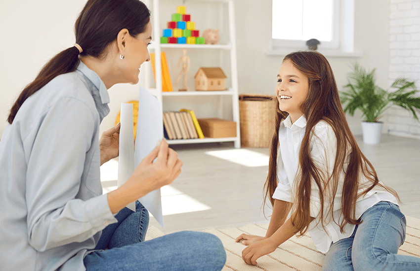 Therapeutin und Mädchen mit langen Haaren sitzen sich in einem freundlich eingerichteten Raum gegenüber, lächeln einander an – Szene eines vertrauensvollen Gesprächs.