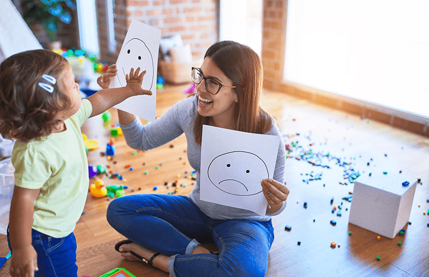 Therapeutin zeigt einem Kleinkind Zeichnungen von Gesichtsausdrücken mit traurigem und neutralem Gesicht, während das Kind die neutrale Zeichnung berührt – Übung zur Emotionswahrnehmung.
